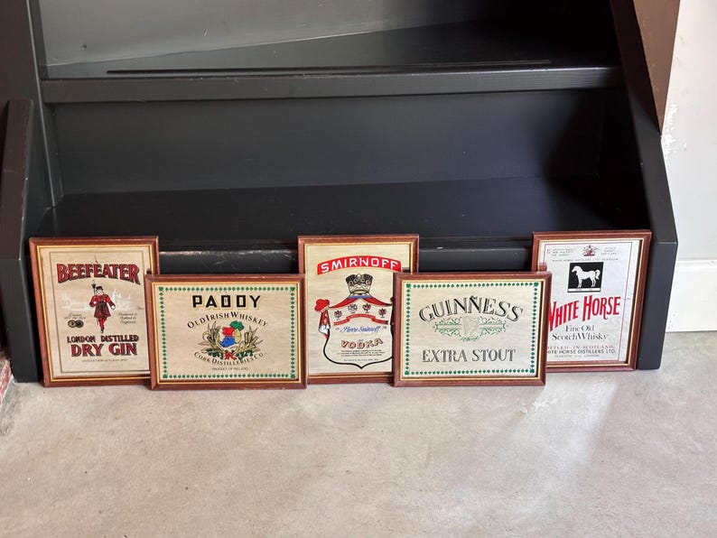 May include: Six framed vintage liquor advertisements, including "Beefeater Dry Gin," "Paddy Old Irish Whiskey," "Smirnoff Vodka," "Guinness Extra Stout," and "White Horse Fine Old Scotch Whisky." Each frame has a brown wooden border.