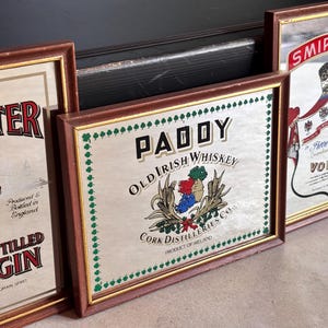 May include: A collection of framed vintage liquor advertisements. The frames are brown with gold trim. The advertisements include "Paddy Old Irish Whiskey", "Beefeater London Distilled Dry Gin", and "Smirnoff Vodka".
