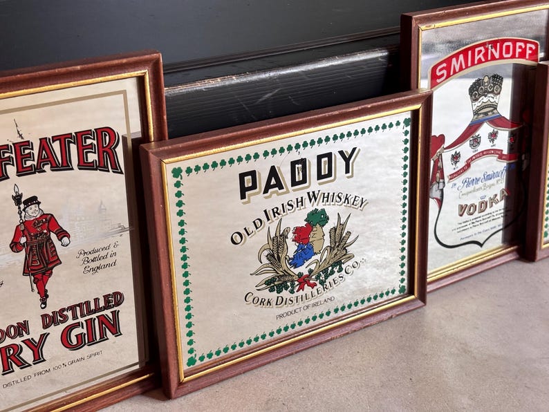 May include: A collection of framed vintage liquor advertisements. The frames are brown with gold trim. The advertisements include "Paddy Old Irish Whiskey", "Beefeater London Distilled Dry Gin", and "Smirnoff Vodka".