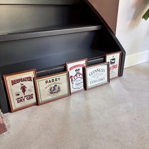 May include: A collection of framed vintage liquor advertisements, including "Beefeater London Distilled Dry Gin," "Paddy Old Irish Whiskey," "Smirnoff Vodka," "Guinness Extra Stout," and "White Horse." The frames are brown, and the images feature various logos and illustrations.