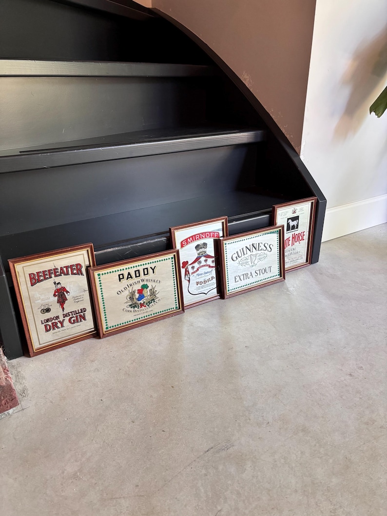 May include: A collection of framed vintage liquor advertisements, including "Beefeater London Distilled Dry Gin," "Paddy Old Irish Whiskey," "Smirnoff Vodka," "Guinness Extra Stout," and "White Horse." The frames are brown, and the images feature various logos and illustrations.