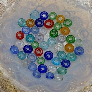 Pode incluir: Uma coleção de contas de vidro coloridas em uma variedade de formas e tamanhos. As contas estão dispostas em um padrão circular sobre uma superfície branca.