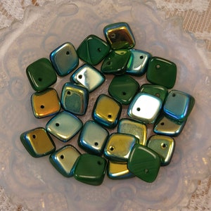 May include: A collection of green and gold iridescent glass square beads with a hole in the center. The beads are arranged in a pile on a white plate.