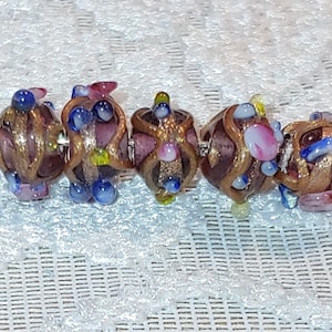 May include: A strand of six glass beads with a metallic gold finish. The beads are decorated with small blue, pink, and yellow glass accents. The beads are strung on a thin wire.