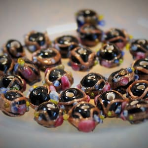 Puede incluir: Una colección de cuentas de vidrio negro con acentos dorados y plateados. Las cuentas están decoradas con purpurina de colores y pequeños trozos de vidrio.
