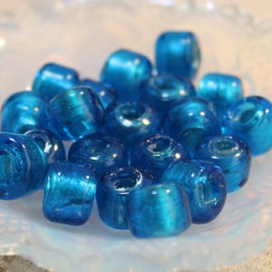 May include: A collection of teal blue glass beads with a metallic sheen. The beads are round and have a hole in the centre for stringing.