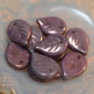 May include: A collection of purple leaf-shaped beads with carved leaf vein details. Each bead has a small hole for stringing. The beads are arranged in a shallow, opalescent dish, showcasing their glossy finish.