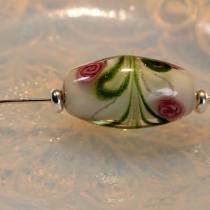 May include: A handcrafted glass bead with a floral design. The bead is oval-shaped, with a white base and pink rose accents, green swirls, and silver detailing. It has a silver pin attached at both ends, perfect for jewelry making.
