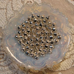 Può includere: Una collezione di piccole, lucenti campanelle d'argento con anelli per infilare, in una ciotola di vetro opalescente decorativa. Le campanelle sono raggruppate, riflettendo la luce, e adatte per lavori artigianali o decorazioni.