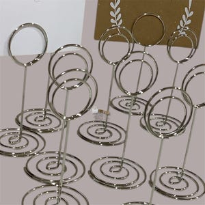 Könnte beinhalten: Eine Sammlung silberfarbener Tischkartenhalter. Jeder Halter hat einen spiralförmigen Fuß, einen schlanken Stiel und eine kreisförmige Oberseite zum Halten von Karten. Anordnung auf neutralem Hintergrund.