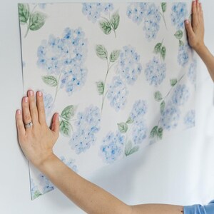 May include: A rectangular sheet of wallpaper featuring a repeating watercolor pattern of light blue hydrangea flower clusters and green leaves on a white background. The floral design is printed in soft, blended shades of blue and green.