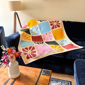 Op de afbeelding: Een handgemaakte gehaakte deken met een raster van kleurrijke vierkanten met bloem- en bladmotieven in tinten geel, blauw, roze, rood en donkergroen. De deken heeft een lichte rand en ligt over een donkerblauwe bank.