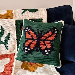 May include: A green crochet pillow with a large butterfly design in orange, black, and white. The pillow is placed on a cream-colored crocheted blanket with green leaf and orange flower patterns. The pillow has a white border.