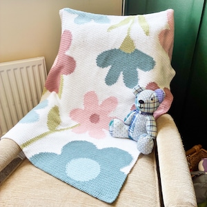Può includere: Una coperta all'uncinetto bianca con un grande motivo floreale nei toni del rosa polvere, verde salvia e blu tenue. Un piccolo orsacchiotto seduto, realizzato con un patchwork di tessuti a quadri e a pois blu, bianchi e grigi, è appoggiato sopra.