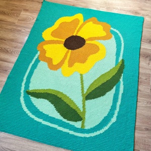 May include: A handmade crochet rug featuring a large yellow flower with a brown center and green leaves. The flower is set against a light blue background within a white oval, all on a turquoise field. The rug is rectangular.