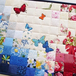 Könnte beinhalten: Ein weißer gesteppter Platzdeckchen mit einem blauen und roten Farbschema. Das Platzdeckchen ist mit verschiedenen Blumenmustern und gestickten Schmetterlingen in Rot-, Blau- und Orangetönen verziert.