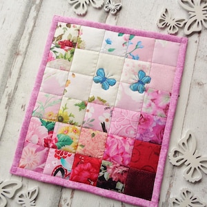 Könnte beinhalten: Eine kleine rosa und weiße Patchwork-Decke mit Blumenmuster und blauen Schmetterlingen. Die Decke besteht aus Quadraten aus verschiedenen Stoffen und ist mit einem rosa Rand eingefasst.