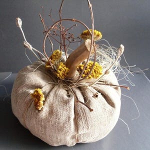 Könnte beinhalten: Ein dekorativer Kürbis aus beigem Stoff, verziert mit einem Holzstiel, getrockneten gelben Blumen und Zweigen. Dieser handgefertigte Artikel eignet sich perfekt für Herbstdekorationen und saisonale Ausstellungen.