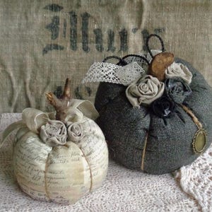 Peut inclure: Deux citrouilles décoratives en tissu. L'une est crème avec une écriture et une ficelle, surmontée d'une brindille et de roses en tissu. L'autre est gris foncé, ornée de roses en tissu, de dentelle et d'un petit pendentif. Les deux sont sur une surface en dentelle.