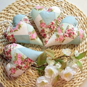 Può includere: Quattro cuscini a forma di cuore fatti a mano con un design patchwork in tessuto floreale nei toni del rosa, bianco e blu. I cuscini sono disposti su una superficie in vimini intrecciato con un rametto di fiori bianchi.