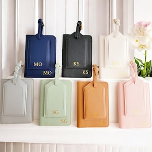 Puede incluir: Una colección de etiquetas de equipaje y portapasaportes personalizados en varios colores, incluyendo azul marino, negro, blanco, gris, verde menta, marrón claro y rosa. Cada etiqueta y portapasaporte tiene iniciales doradas.
