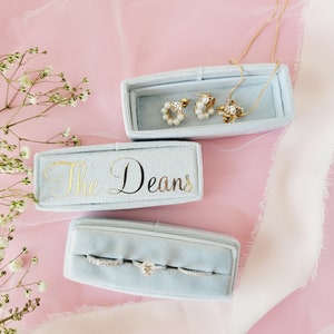 Puede incluir: Tres cajas de anillos de terciopelo azul claro con letras doradas. La caja superior dice "The Deans". La caja del medio contiene tres anillos. La caja inferior contiene un collar y un par de pendientes.
