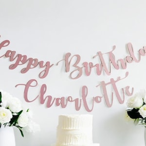 Puede incluir: Una pancarta rosa y blanca con el texto "Happy Birthday Charlotte" colgada en una pared blanca. La pancarta está hecha de papel y tiene un acabado brillante. Hay un pastel blanco delante de la pancarta.