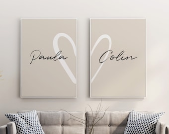 Op de afbeelding: Twee beige canvas prints met witte hartvormen en de namen "Paula" en "Colin" in zwarte schrijfletters. De prints hangen aan een witte muur boven een grijze bank met blauwe, gepatroneerde kussens.