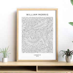 May include: A framed print featuring a gray and white floral pattern, labeled "Larkspur 1875" by William Morris. The artwork is displayed on a light wood desk, with green plants in the foreground.