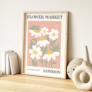 May include: A framed print with a pink background and white daisies with yellow centers. The print says "FLOWER MARKET" at the top and "THE FLORAL GALLERY LONDON" at the bottom.
