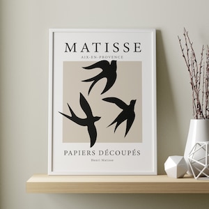 May include: A framed art print featuring a beige background with three black bird silhouettes. The print is titled "MATISSE PAPIERS D&Eacute;COUP&Eacute;S" and is displayed on a wooden shelf with decorative items, including a white vase and geometric shapes.