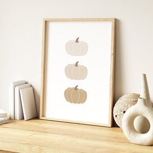May include: A framed print with a minimalist design featuring three pumpkins in shades of beige and brown. The pumpkins are stacked vertically in the centre of the print.