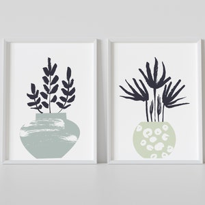 May include: Two framed prints of abstract botanical illustrations. The prints feature stylised plants in green and white vases with black outlines. The prints are set against a white background.
