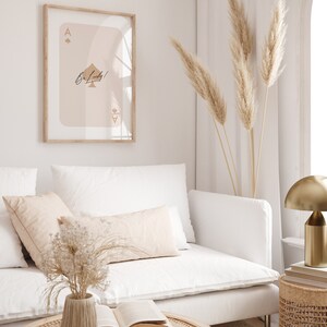 May include: A white couch with a beige throw pillow and a vase of dried flowers on a coffee table. A framed print of an ace of spades with the text "Be Lucky!" hangs on the wall behind the couch. A gold lamp and a wicker side table are also visible in the image.