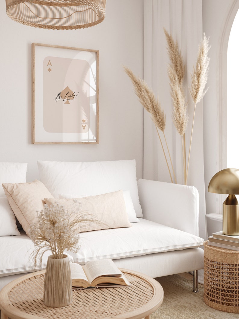 May include: A white couch with a beige throw pillow and a vase of dried flowers on a coffee table. A framed print of an ace of spades with the text "Be Lucky!" hangs on the wall behind the couch. A gold lamp and a wicker side table are also visible in the image.