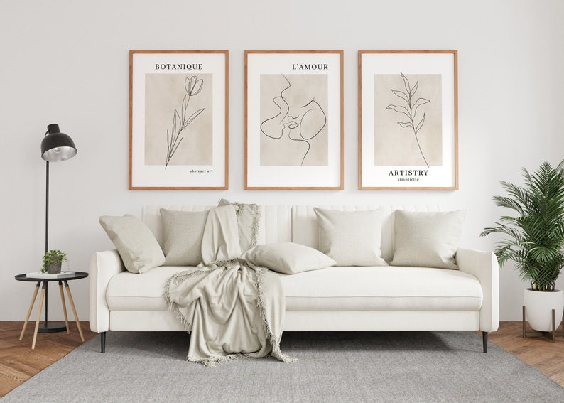 K&ouml;nnte beinhalten: Drei minimalistische Line-Art-Drucke mit Holzrahmen h&auml;ngen an einer wei&szlig;en Wand &uuml;ber einem wei&szlig;en Sofa mit beigen Kissen und einer beigen Decke. Die Drucke zeigen eine Blume, ein Gesicht und ein Blatt. Die Drucke sind mit "Botanique", "L'Amour" und "Artistry" betitelt.