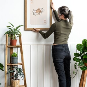 Puede incluir: Una mujer cuelga una impresi&oacute;n enmarcada en una pared blanca. La impresi&oacute;n presenta un as de picas estilizado con el texto "No se trata de las cartas que te reparten, sino de c&oacute;mo las juegas."