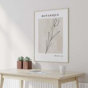 K&ouml;nnte beinhalten: Ein gerahmter Druck mit einer Strichzeichnung einer Blume auf beigefarbenem Hintergrund. Der Druck tr&auml;gt den Text "BOTANIQUE" und "abstrakte Kunst". Der Druck h&auml;ngt an einer wei&szlig;en Wand &uuml;ber einem Holztisch mit zwei Sukkulenten in T&ouml;pfen und einer wei&szlig;en Tasse.
