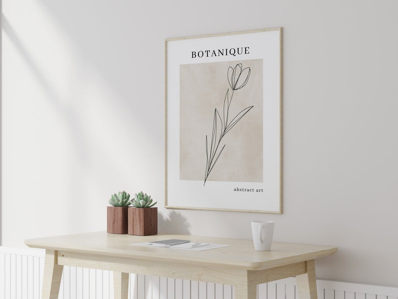 K&ouml;nnte beinhalten: Ein gerahmter Druck mit einer Strichzeichnung einer Blume auf beigefarbenem Hintergrund. Der Druck tr&auml;gt den Text "BOTANIQUE" und "abstrakte Kunst". Der Druck h&auml;ngt an einer wei&szlig;en Wand &uuml;ber einem Holztisch mit zwei Sukkulenten in T&ouml;pfen und einer wei&szlig;en Tasse.
