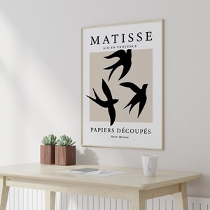 May include: A framed art print featuring a minimalist design with three black bird silhouettes against a beige background. The print includes the text "MATISSE AIX-EN-PROVENCE PAPIERS D&Eacute;COUP&Eacute;S Henri Matisse". The artwork is displayed on a light-colored wooden desk.
