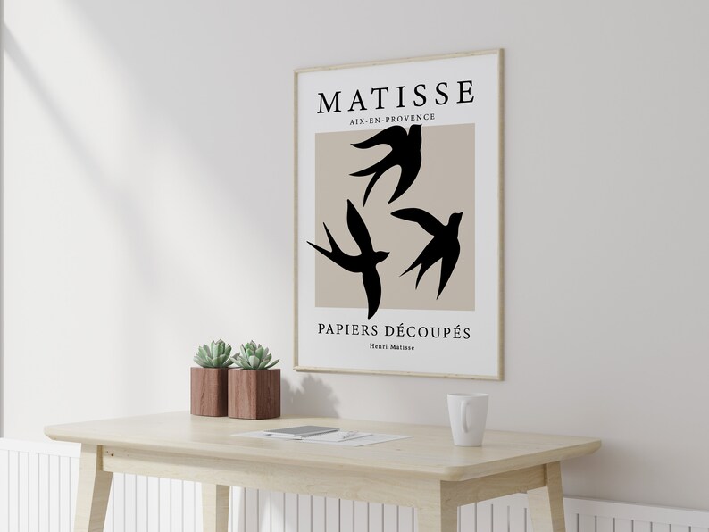 May include: A framed art print featuring a minimalist design with three black bird silhouettes against a beige background. The print includes the text "MATISSE AIX-EN-PROVENCE PAPIERS D&Eacute;COUP&Eacute;S Henri Matisse". The artwork is displayed on a light-colored wooden desk.