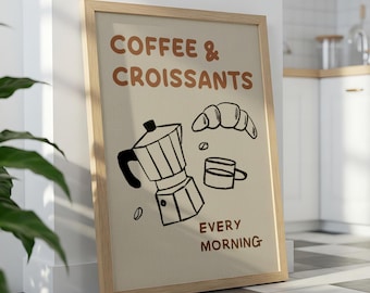 May include: A framed print with the words "Coffee & Croissants EVERY MORNING" in brown lettering. The print features a black and white illustration of a coffee pot, croissant, coffee cup, and coffee bean. The frame is made of light-coloured wood.