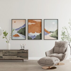 Pode incluir: Tr&ecirc;s impress&otilde;es de aquarela de paisagens emolduradas em um estilo minimalista, representando montanhas, &aacute;gua e uma lua ou sol. As impress&otilde;es est&atilde;o penduradas em uma parede branca acima de uma mesa de console de madeira e uma poltrona cinza claro com um pufe combinando.