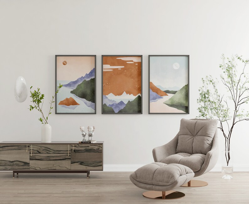 Pode incluir: Tr&ecirc;s impress&otilde;es de aquarela de paisagens emolduradas em um estilo minimalista, representando montanhas, &aacute;gua e uma lua ou sol. As impress&otilde;es est&atilde;o penduradas em uma parede branca acima de uma mesa de console de madeira e uma poltrona cinza claro com um pufe combinando.