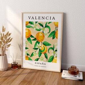 Op de afbeelding: Een ingelijst afdruk met een aquarel-illustratie van sinaasappelbloesems en sinaasappelen. De afdruk heeft de tekst "VALENCIA" bovenaan en "ESPA&Ntilde;A TIERRA DE NARANJAS" onderaan.