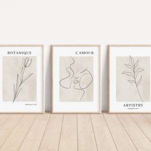 K&ouml;nnte beinhalten: Drei minimalistische Line-Art-Drucke in Holzrahmen. Der erste Druck zeigt eine einzelne Blume mit dem Text "BOTANIQUE abstract art". Der zweite Druck zeigt die Gesichter eines Paares mit dem Text "L'AMOUR". Der dritte Druck zeigt ein einzelnes Blatt mit dem Text "ARTISTRY simplicit&eacute;".
