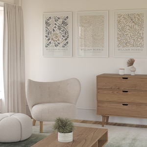 May include: Three framed prints with botanical illustrations and the text "William Morris" hanging on a white wall above a wooden dresser with three drawers. A white armchair and a white ottoman are in the foreground.