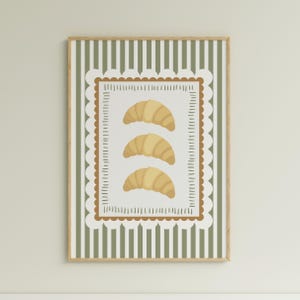 May include: A framed print depicting three illustrated croissants. The artwork has a white background with a brown scalloped border and a green striped background. The frame is a light wood colour.