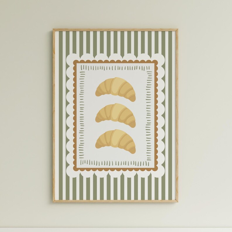 May include: A framed print depicting three illustrated croissants. The artwork has a white background with a brown scalloped border and a green striped background. The frame is a light wood colour.