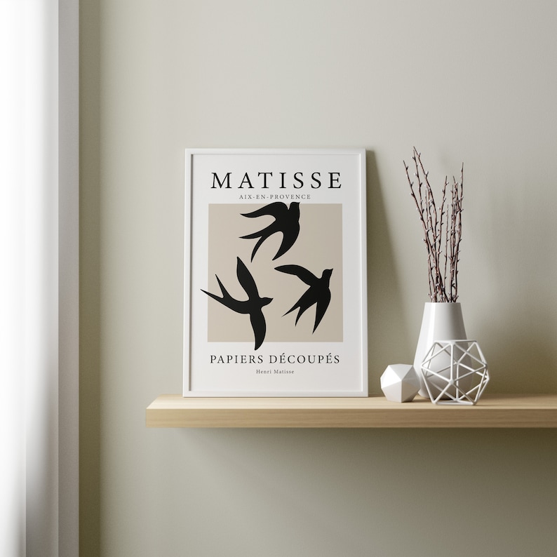 May include: A framed art print featuring a beige background with three black bird silhouettes. The print is titled "MATISSE PAPIERS D&Eacute;COUP&Eacute;S" and is displayed on a wooden shelf with decorative items, including a white vase and geometric shapes.